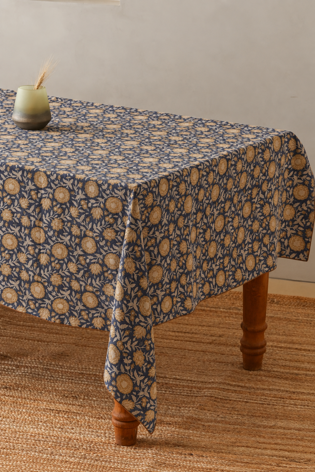 A rectangular tablecloth with a navy blue and beige floral block-print pattern draped over a wooden table, set on a woven jute rug against a neutral wall.