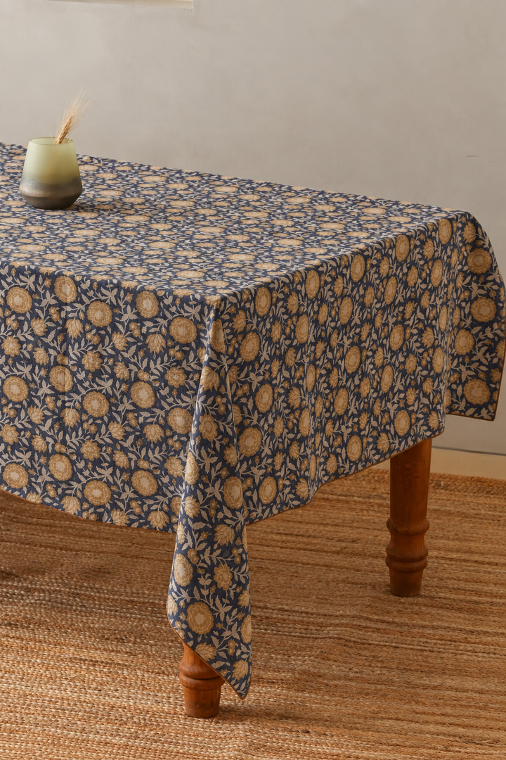 A rectangular tablecloth with a navy blue and beige floral block-print pattern draped over a wooden table, set on a woven jute rug against a neutral wall.