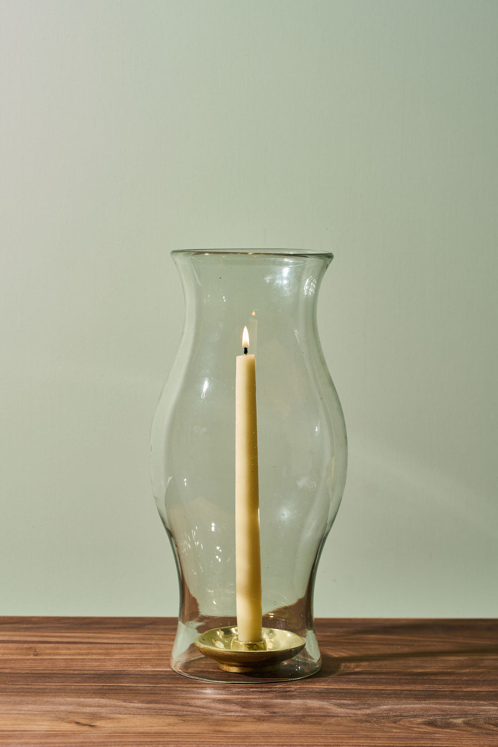 A clear glass vase with a flared rim and a single cream candle standing inside on a small gold base, placed on a wooden surface against a pale green wall.