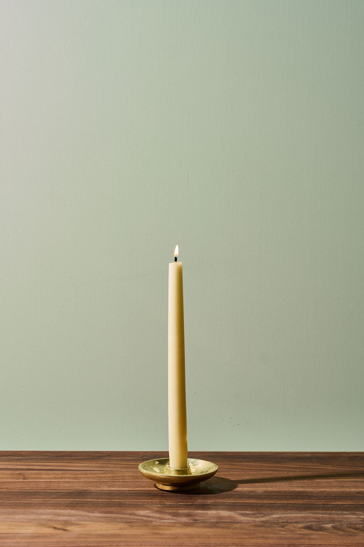 A single lit cream-colored taper candle standing upright in a small round metallic holder on a wooden surface against a pale sage green wall.