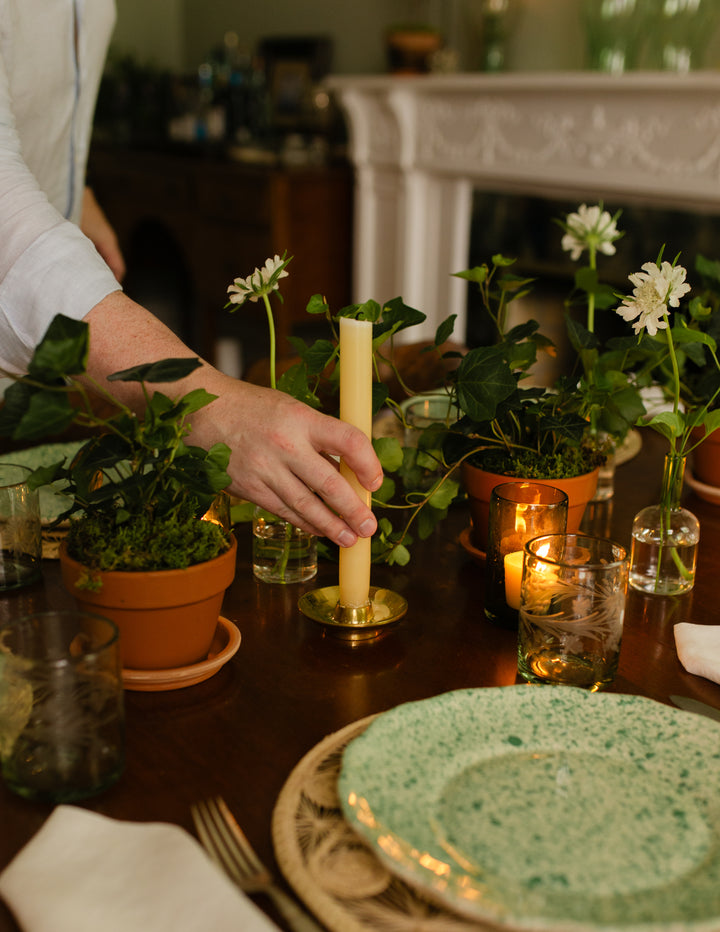 A hand adjusts a lit cream pillar candle in a brass holder on a dark wooden dining table decorated with potted green plants, white flowers, and lit tea lights. A green and white speckled ceramic plate is visible in the foreground.