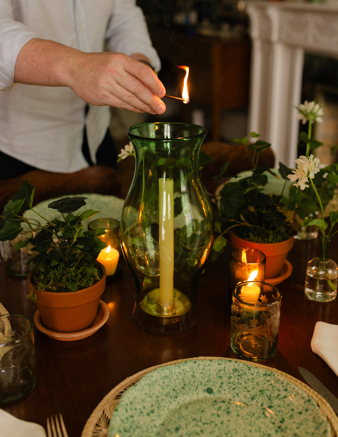 A green glass vase candle holder with a lit taper candle on a dark wooden table, surrounded by potted green plants, small glass candle holders with flames, and a person's hand lighting a candle in the background.
