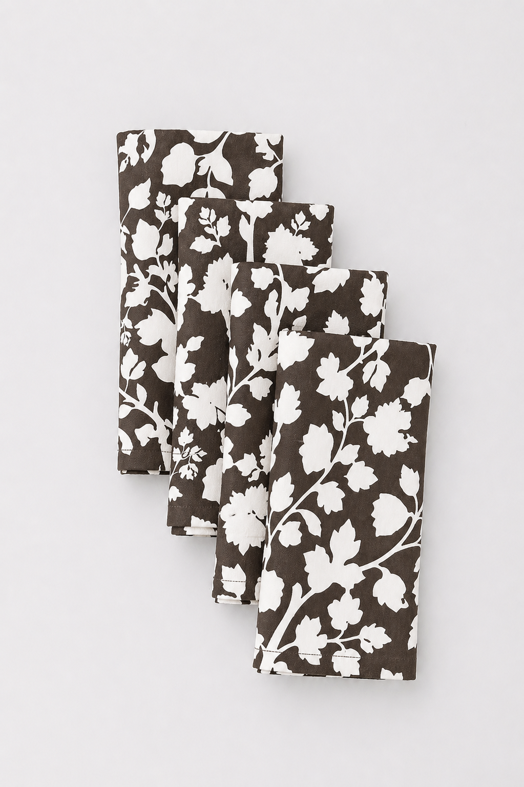 Four folded rectangular fabric napkins with a white floral vine pattern on a dark brown background, arranged in a staggered stack on a light gray background.