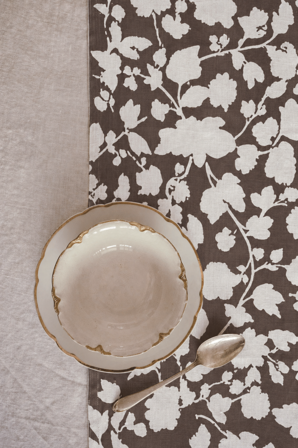 An off-white ceramic bowl with a scalloped rim and gold detailing, placed on a brown and white floral patterned tablecloth next to a silver spoon.