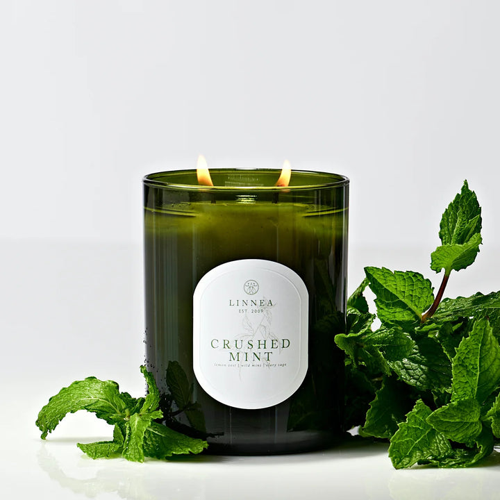 A lit Linnea 'Crushed Mint' scented candle in a dark green glass jar, styled with fresh mint leaves on a clean white background.