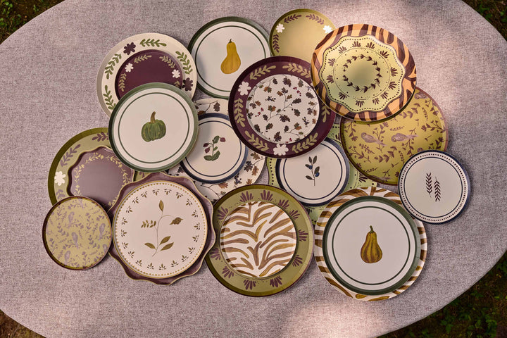 A collection of decorative  plates with autumn-themed patterns, including pumpkins, pears, leaves, and floral motifs, arranged on a round gray table.