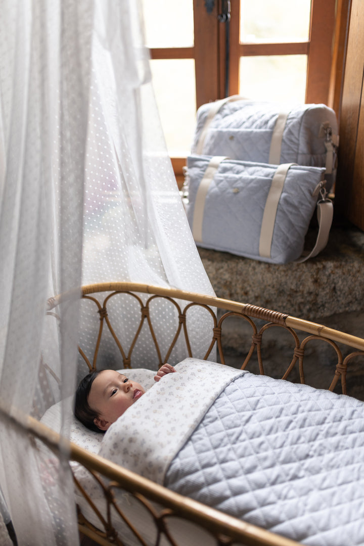 A baby rests in a woven bassinet under a white canopy, wrapped in a light blue and white quilted blanket. Matching light blue and white travel bags are stacked on a stone surface near a window in the background.