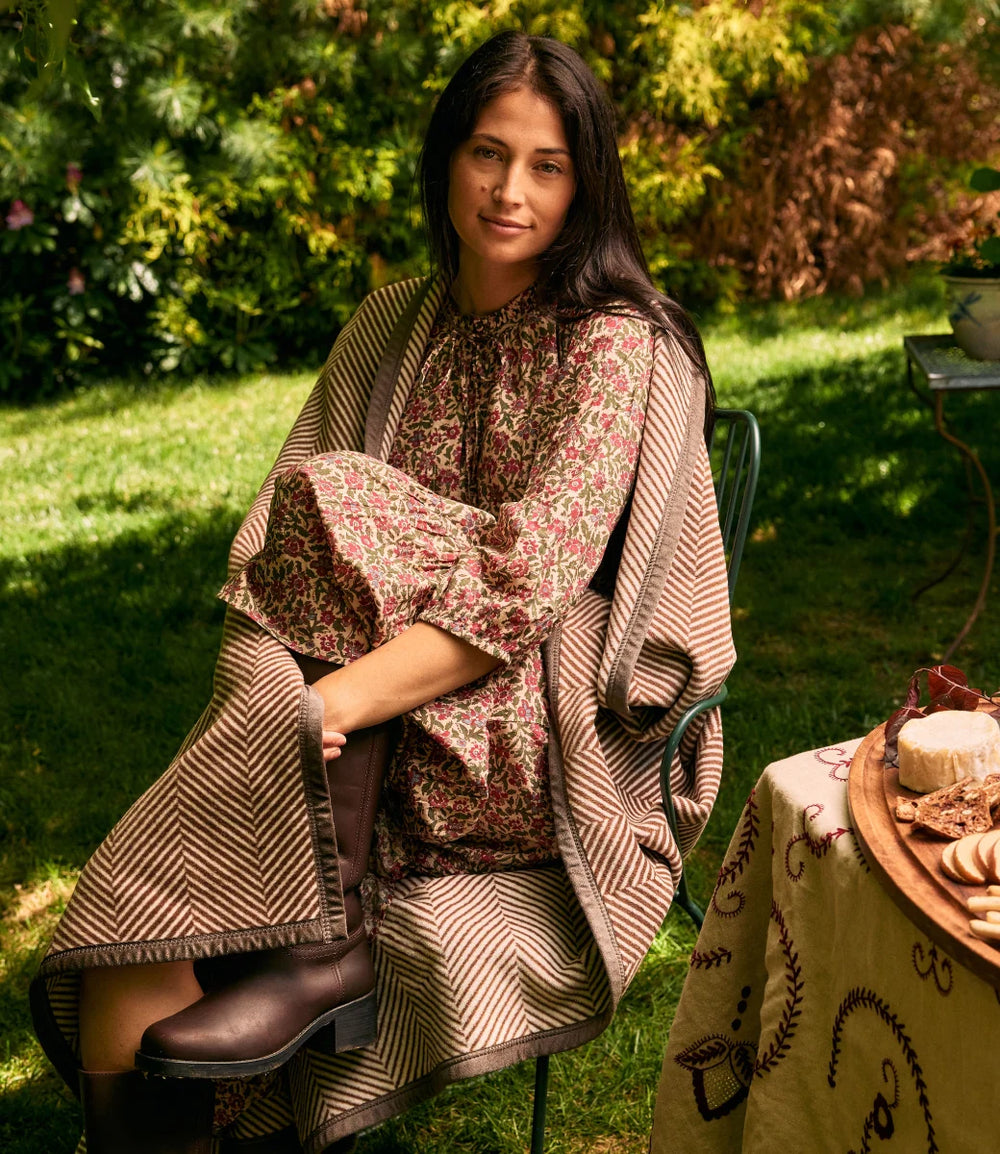 A woman sits outdoors on a garden lawn wearing a patterned bohemian-style dress with a large striped knit shawl draped over her shoulders. She is wearing dark brown leather boots, with a table featuring bread and pastries visible in the foreground.