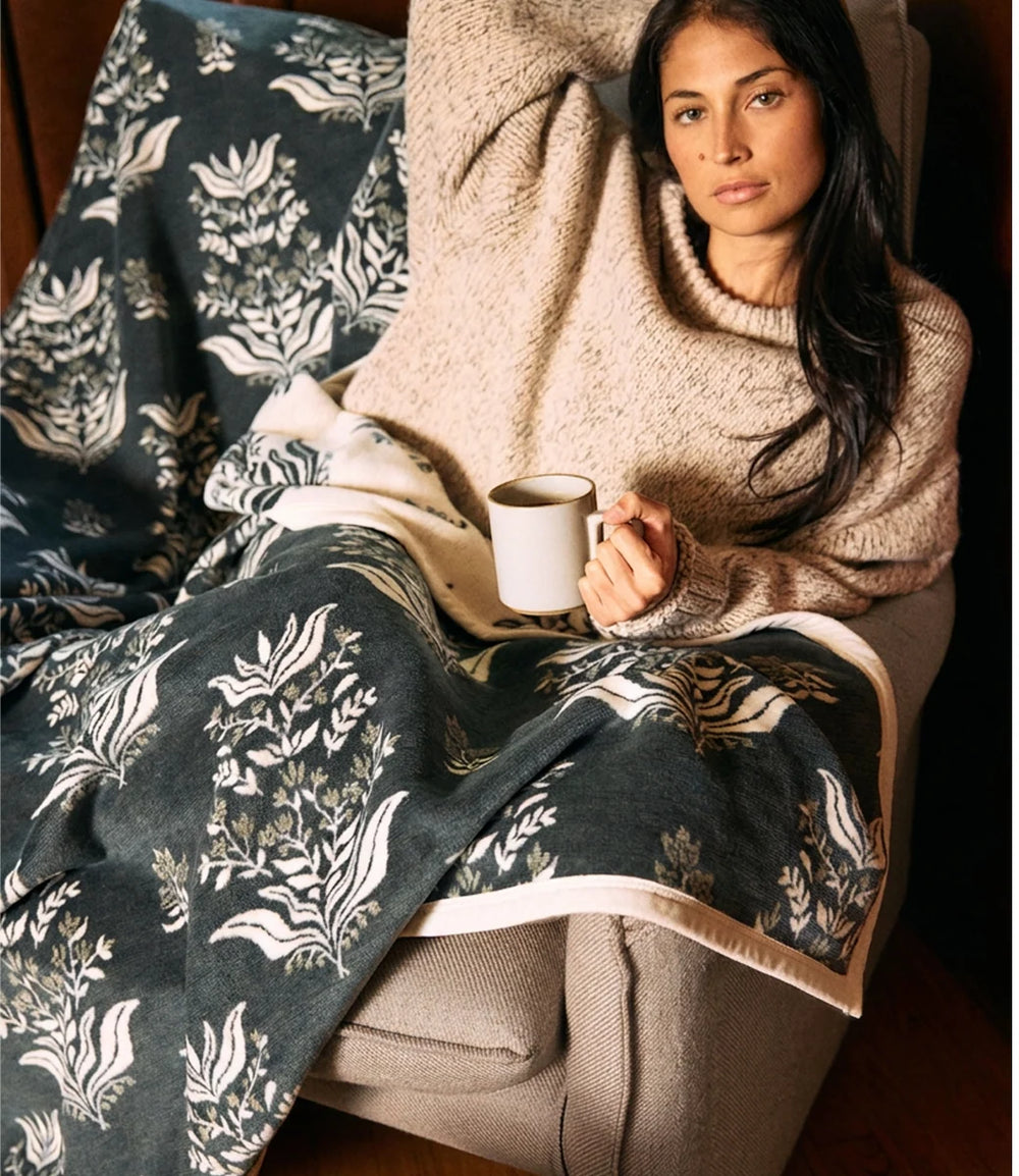 A woman reclining in a chair wrapped in a cozy beige knit sweater and covered with a dark teal floral blanket, holding a white mug.