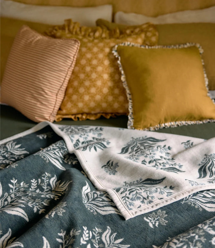 A styled bedding scene with multiple decorative pillows and a folded patterned throw blanket in muted teal, cream, and warm yellow tones.