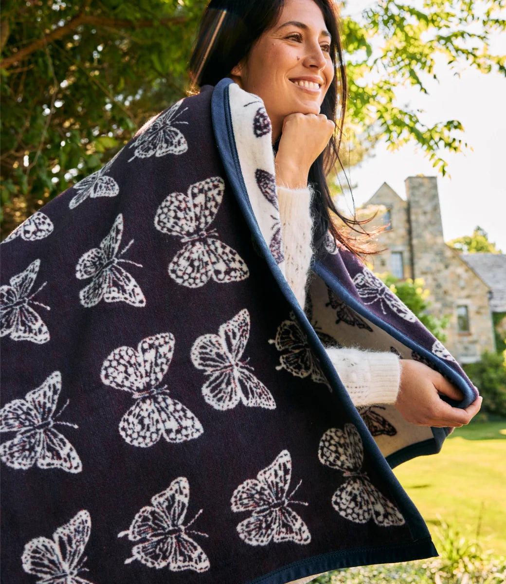 A woman outdoors wrapped in a dark navy blanket with a white butterfly pattern and a cream lining, wearing a white knit sweater. Trees and a stone house are visible in the background.