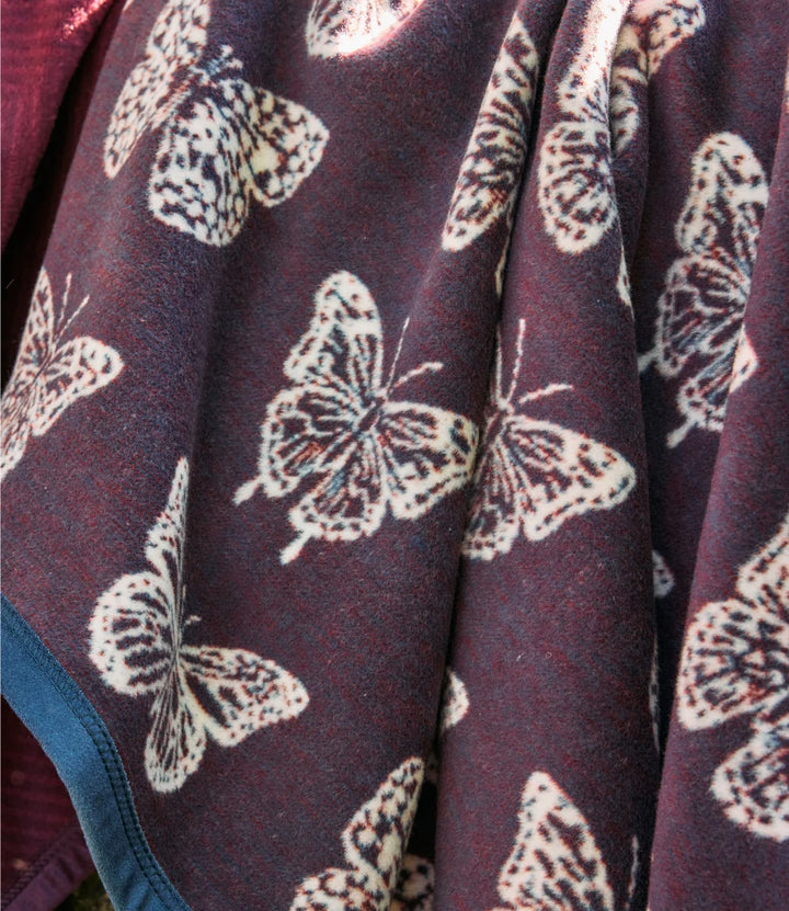 A close-up of a dark plum-purple fleece blanket with a repeating cream-colored butterfly pattern and a blue border trim.