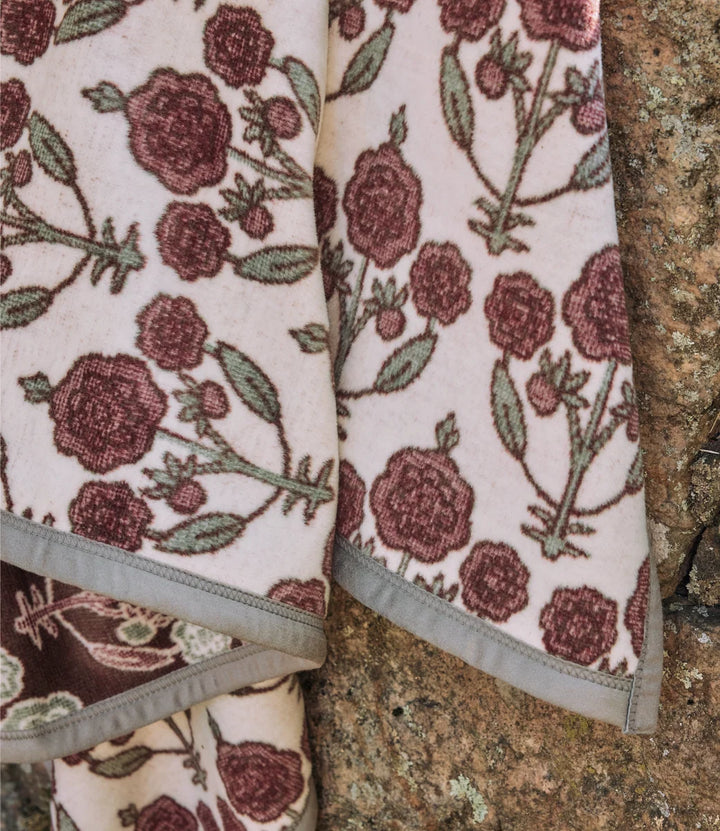 A close-up of a woven textile with a floral pattern in muted burgundy and olive green on a warm cream background, folded with a gray stitched border, placed against a rough stone surface.