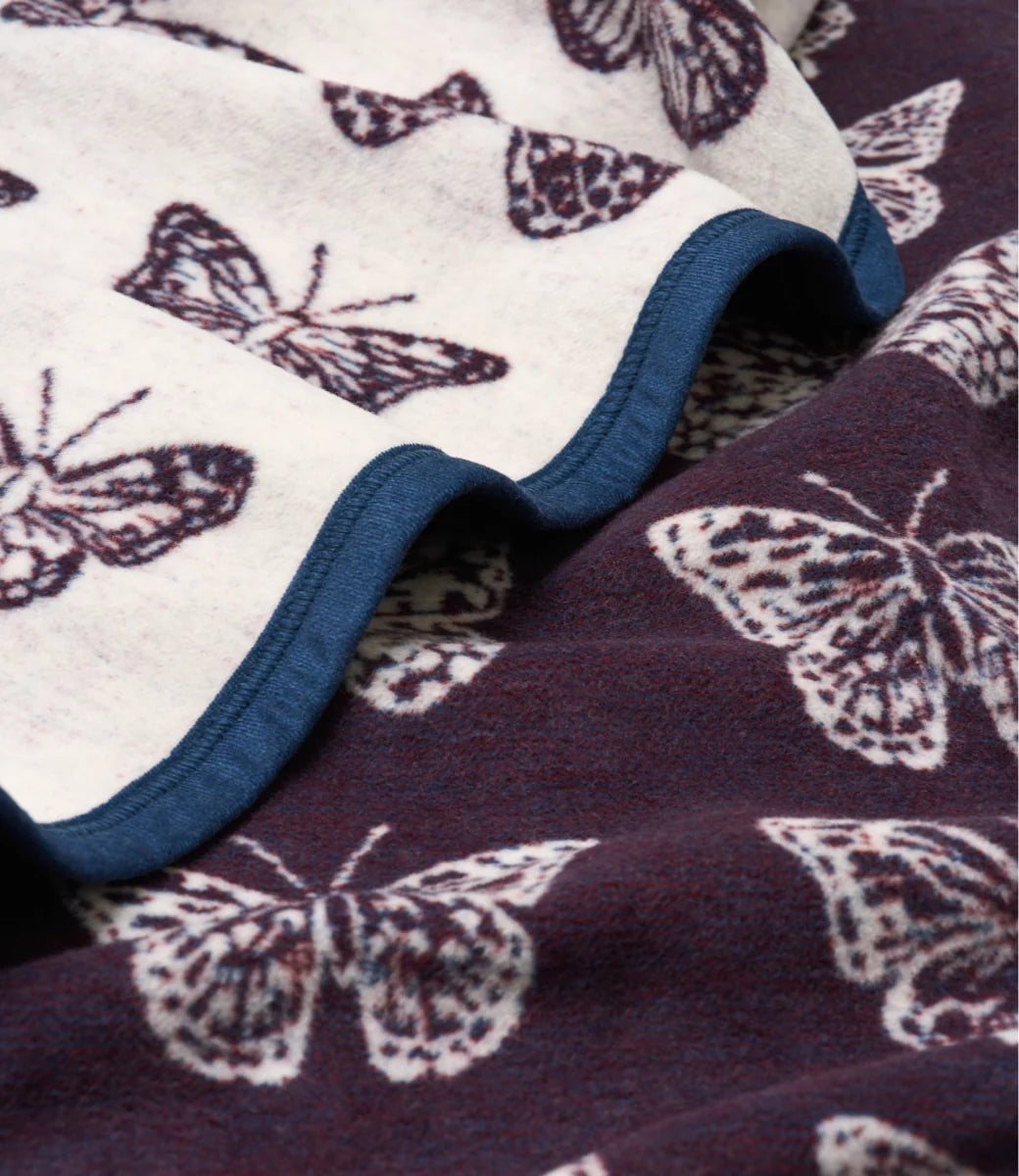 A close-up of a soft fleece blanket with a butterfly pattern. The top section is cream-colored with dark brown butterfly prints and a blue ruffled edge, while the bottom section is dark plum-purple with light cream butterfly prints.