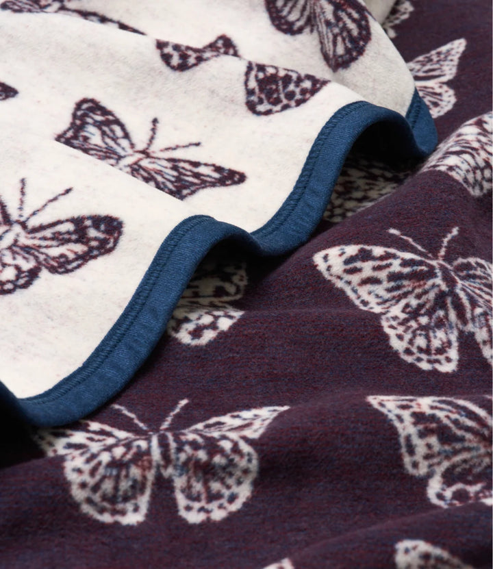 A close-up of a soft fleece blanket with a butterfly pattern. The top section is cream-colored with dark brown butterfly prints and a blue ruffled edge, while the bottom section is dark plum-purple with light cream butterfly prints.