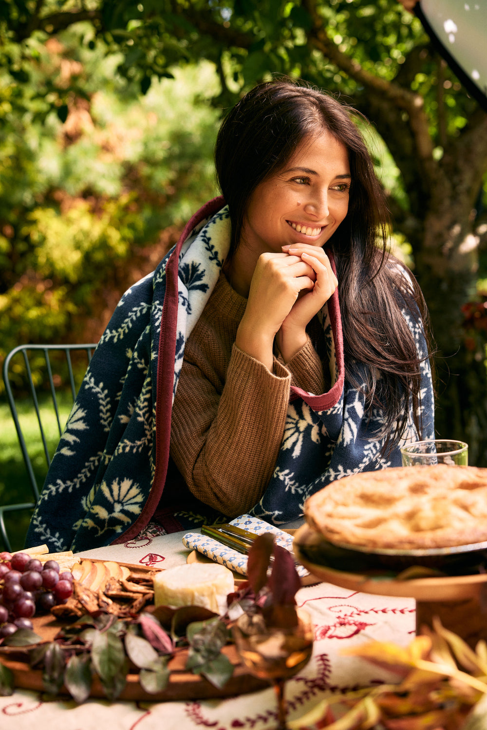 A woman smiling at an outdoor table with a pie, grapes, cheese, and dried leaves, wrapped in a navy blue floral blanket over a brown sweater.