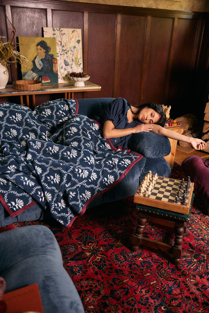 A woman rests on a blue sofa in a cozy, wood-paneled room, covered with a navy and white patterned knit blanket. A small wooden chessboard sits on a table nearby, and a red patterned rug covers the floor.