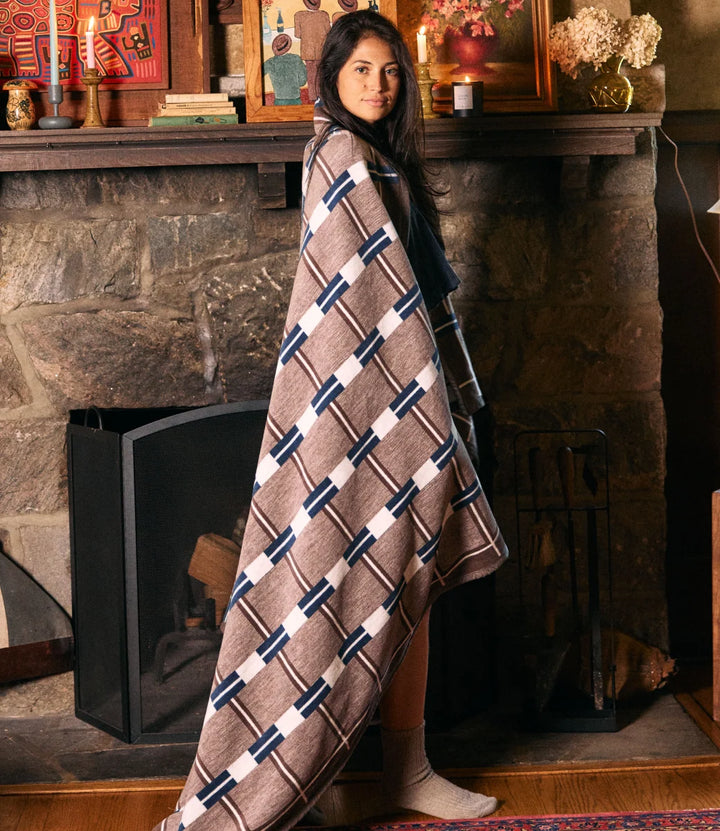 A woman wrapped in a brown patterned blanket standing in front of a rustic stone fireplace.