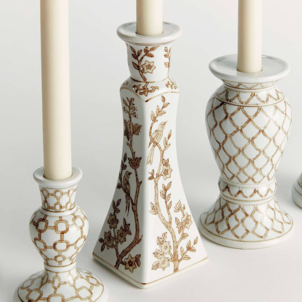 Three white ceramic candlestick holders with gold-toned geometric and floral patterns, each holding a cream-colored candle, arranged on a light neutral background.