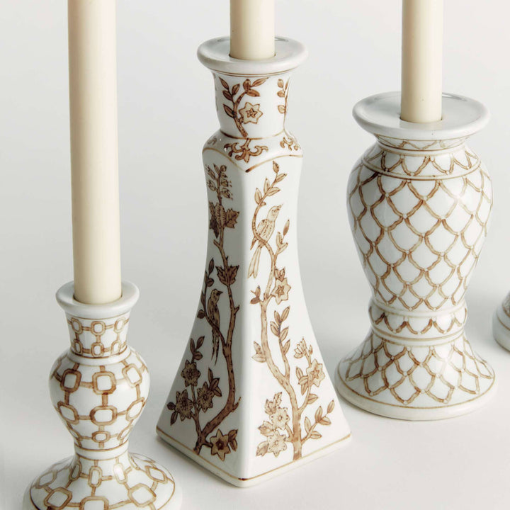 Three white ceramic candlestick holders with gold-toned geometric and floral patterns, each holding a cream-colored candle, arranged on a light neutral background.