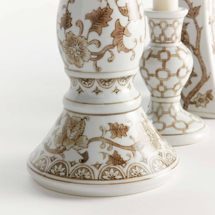 Close-up of a white ceramic candlestick holder with brown floral and scroll patterns, with two additional holders blurred in the background.
