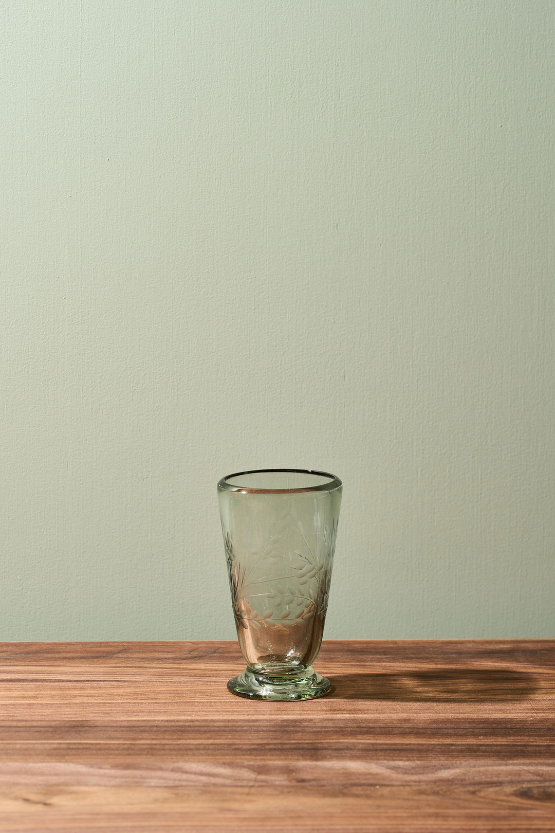 A small clear glass tumbler with a subtle etched floral pattern and a thin black rim, sitting on a wooden tabletop against a light sage green wall.