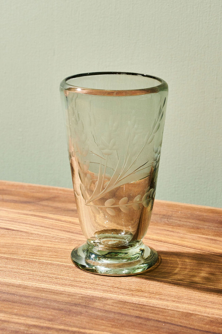 A clear glass tumbler with a footed base and subtle etched floral detailing, standing on a wooden tabletop against a pale green wall.