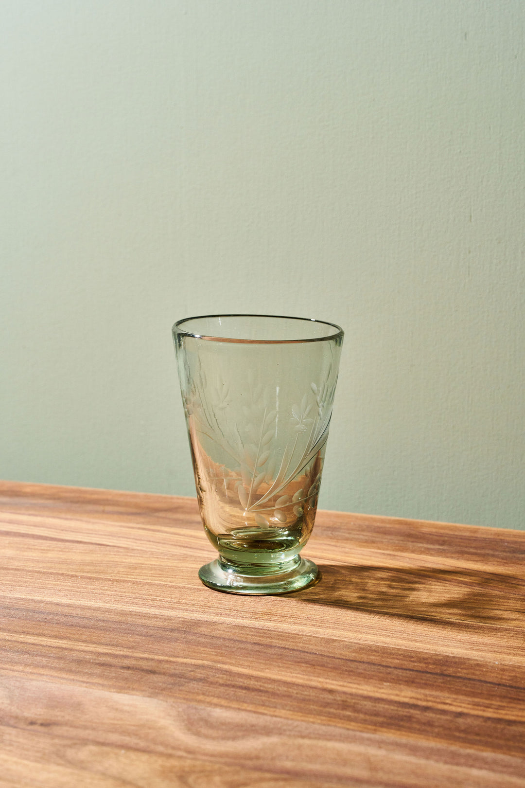 A clear glass tumbler with etched floral patterns and a small footed base, placed on a wooden tabletop against a pale green wall.