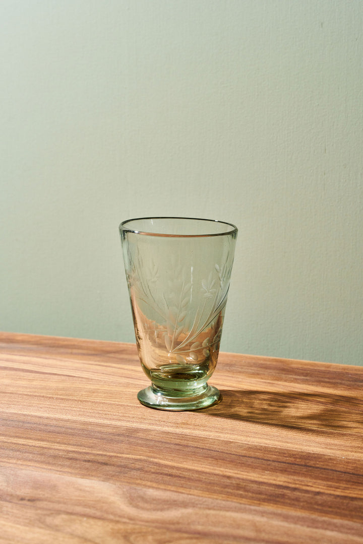 A clear glass tumbler with etched floral patterns and a small footed base, placed on a wooden tabletop against a pale green wall.