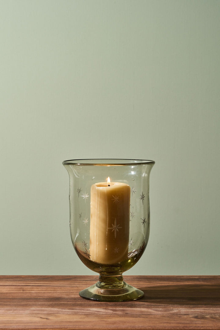 A lit cream-colored pillar candle inside a clear amber glass hurricane lantern with etched star patterns, set on a wooden surface against a muted sage green background.