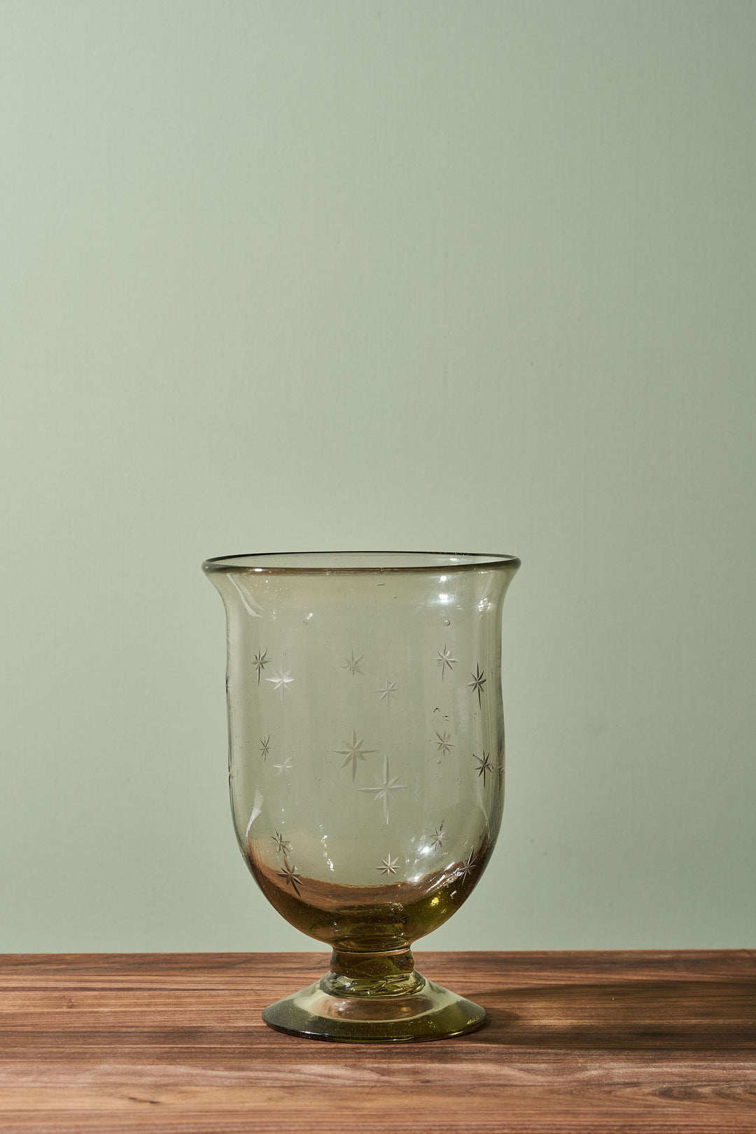A clear glass candle holder with a footed base and etched star patterns, resting on a wooden surface against a sage green background.