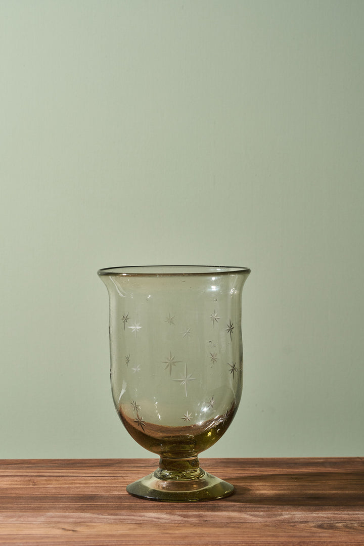 A clear glass candle holder with a footed base and etched star patterns, resting on a wooden surface against a sage green background.