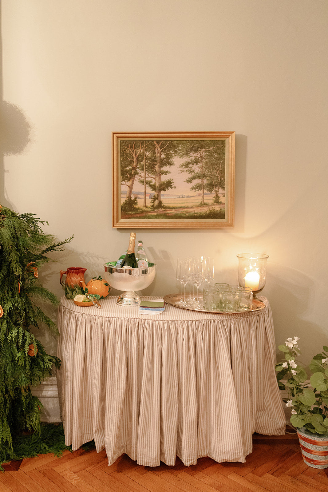 A styled interior scene with a round table covered in a beige vertically striped ruffled tablecloth, holding glassware, a candle, and small decor items. Above the table hangs a framed landscape painting on a warm beige wall, with plants and wood flooring visible around the scene.