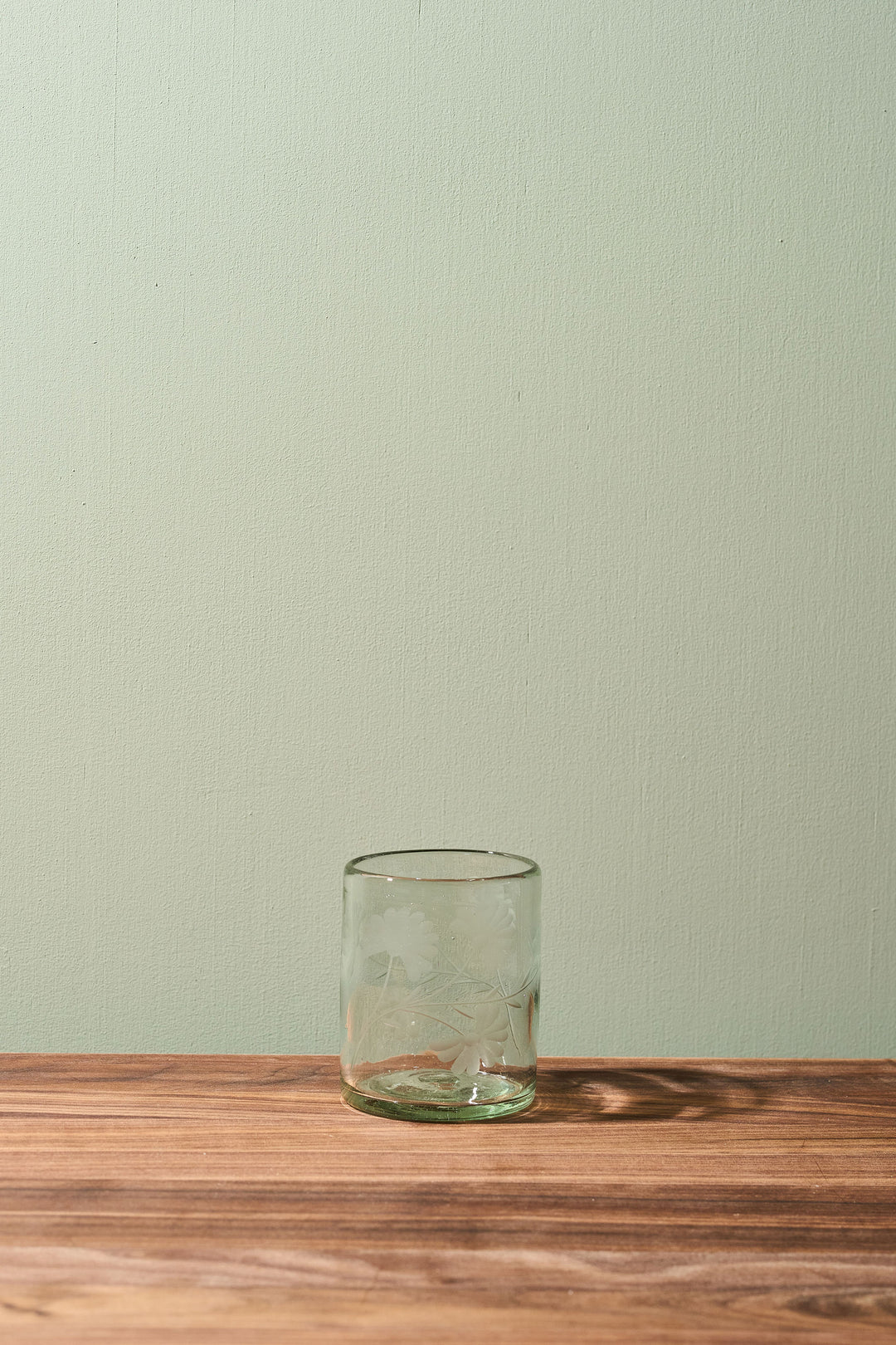 A clear glass tumbler with a subtle embossed floral pattern, resting on a wooden tabletop against a light sage green wall.