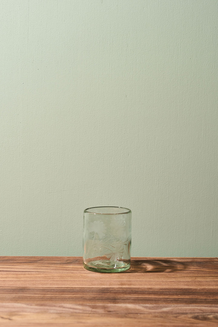 A clear glass tumbler with a subtle embossed floral pattern, resting on a wooden tabletop against a light sage green wall.
