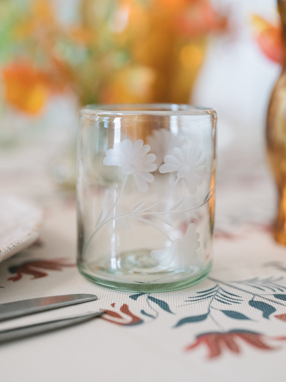 A clear glass candle holder with a frosted white floral design, set on a patterned table runner with blurred dining table elements in the background.