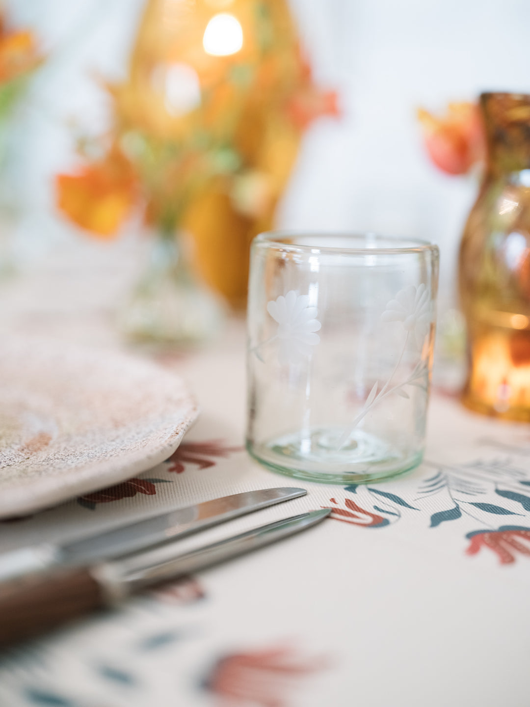 A clear glass candle holder with delicate white floral etched details, styled on a patterned table runner alongside a knife and a warm, softly blurred amber bottle in the background.