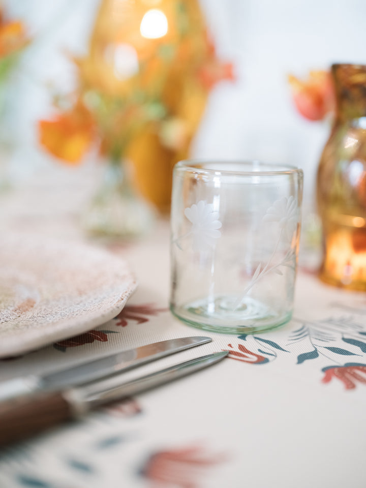 A clear glass candle holder with delicate white floral etched details, styled on a patterned table runner alongside a knife and a warm, softly blurred amber bottle in the background.