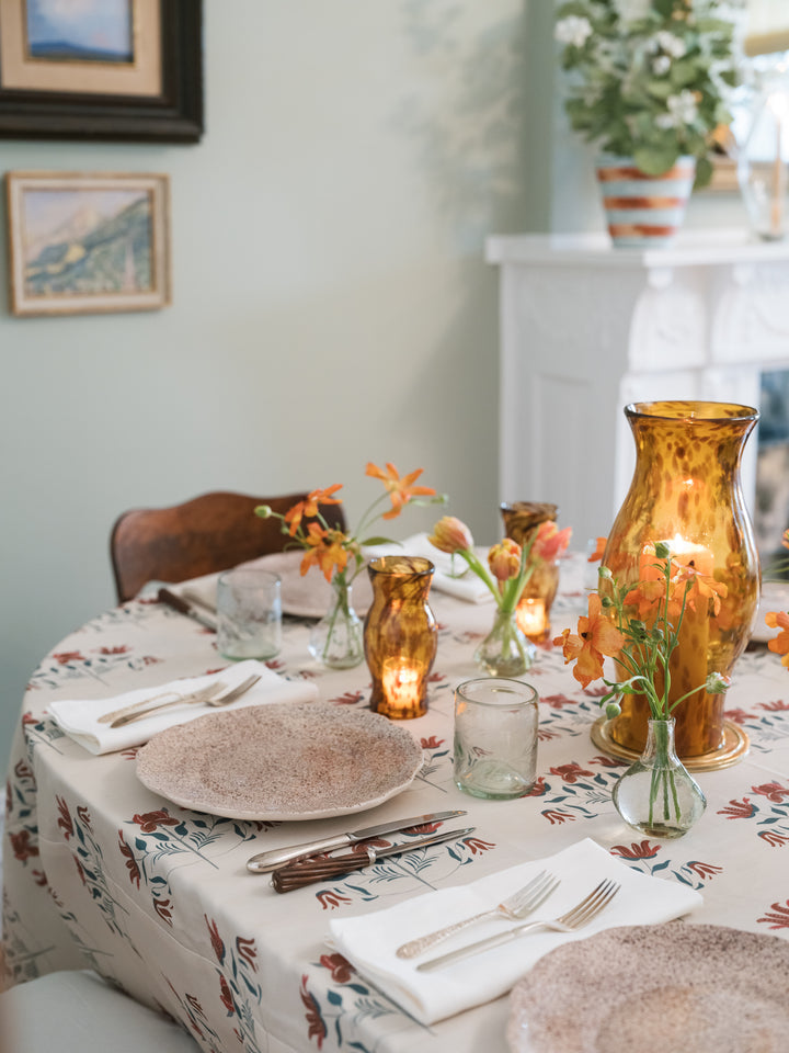 A styled dining table in a home setting, featuring a beige tablecloth with a muted floral pattern, textured beige plates, white napkins with silver cutlery, and amber glass candle vases and small floral arrangements.