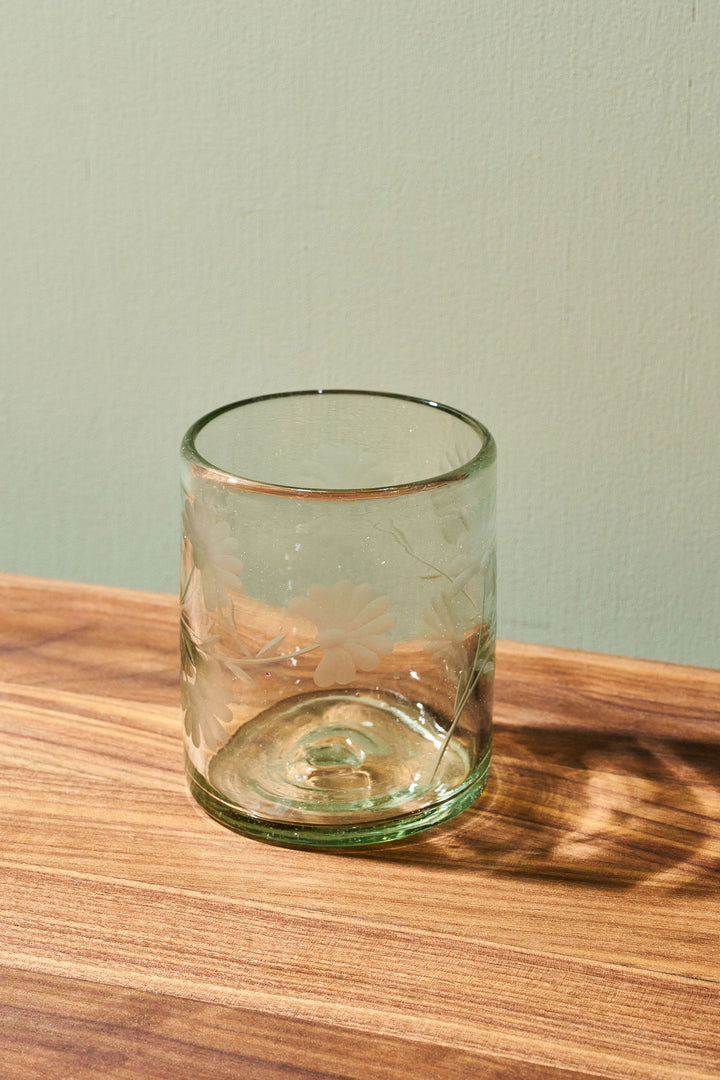 A clear green-tinted glass tumbler with a subtle floral etched pattern, sitting on a wooden tabletop against a light sage green wall.