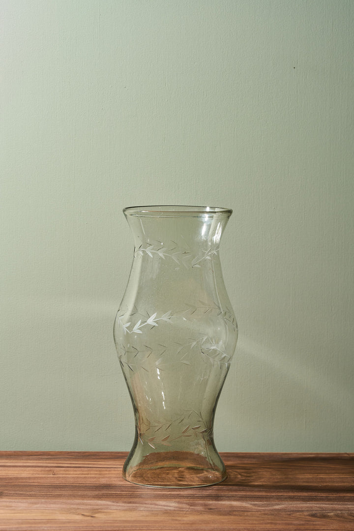 A clear glass vase with a flared rim and etched geometric leaf patterns, standing on a wooden surface against a light sage green wall.