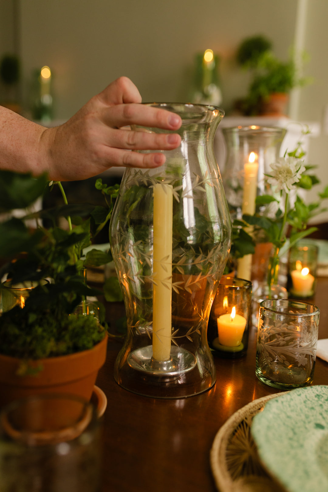 A hand holds a tall, clear glass vase with etched leaf patterns containing a lit yellow taper candle, set on a warmly lit wooden table surrounded by potted plants and other candles.