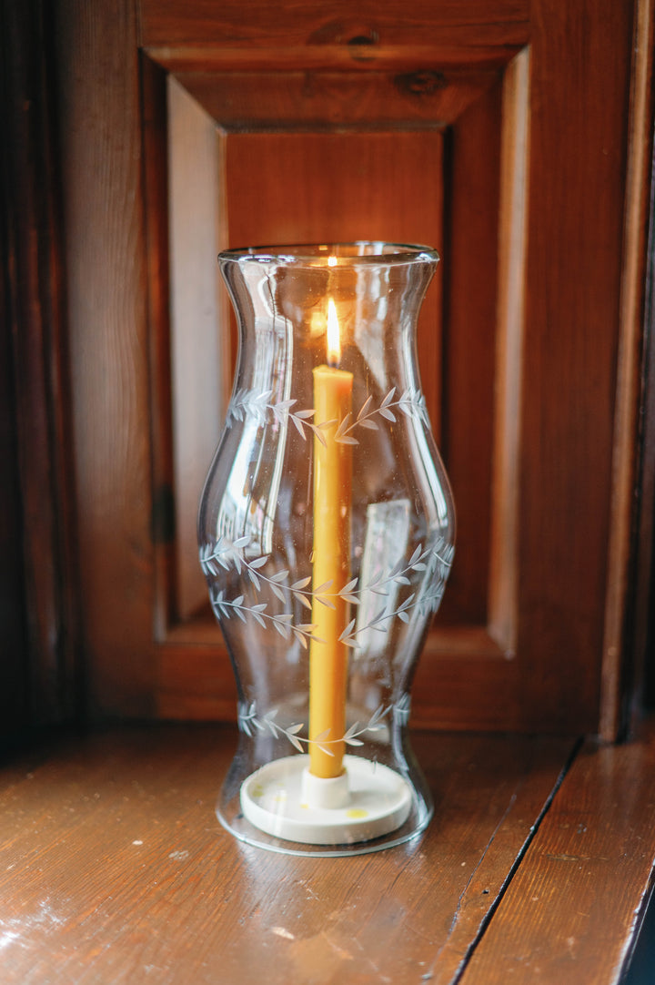 A clear glass hurricane lantern with etched leaf patterns, holding a lit yellow taper candle on a wooden surface in front of a dark wood paneled background.