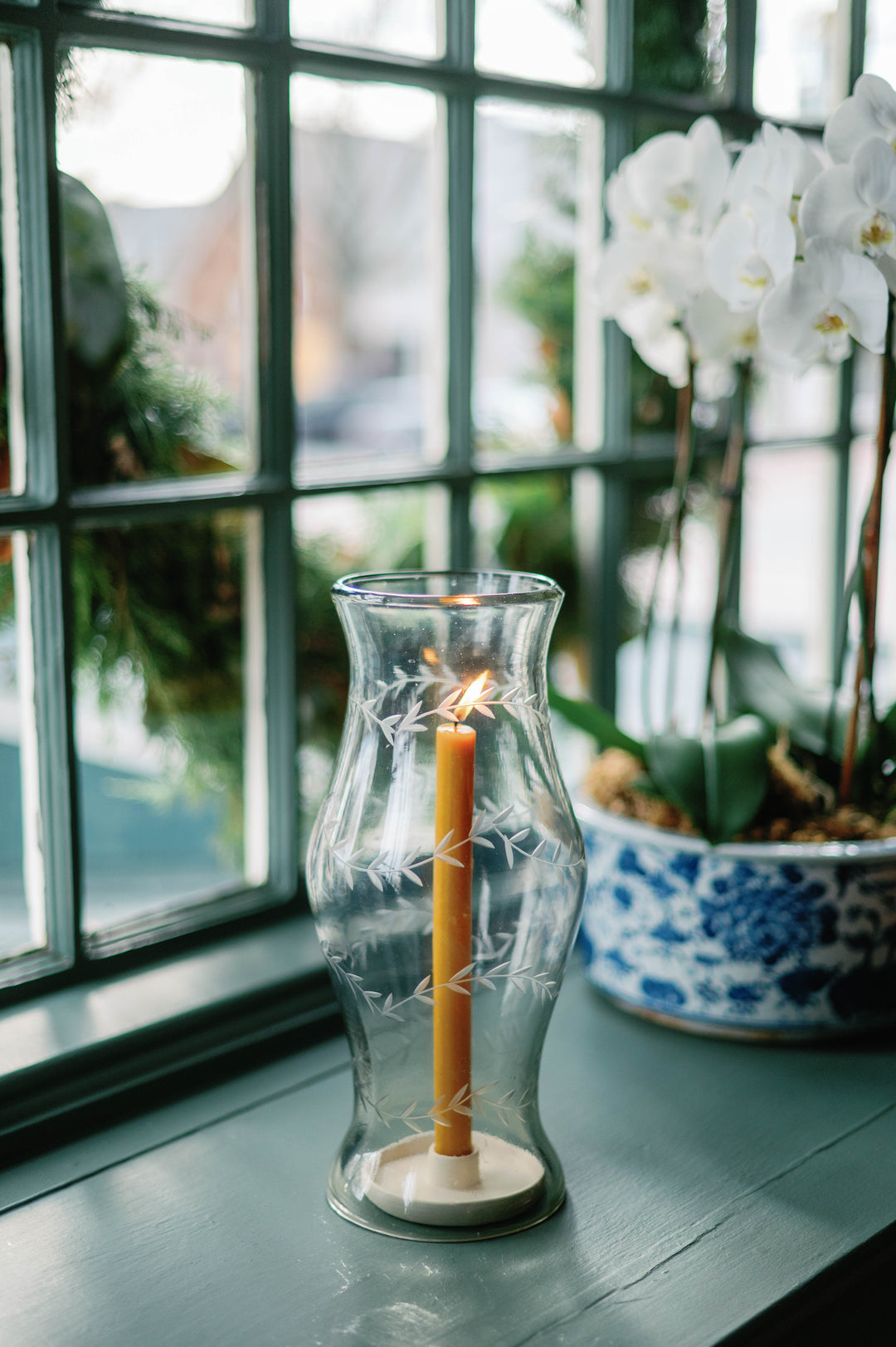 A clear glass candle holder with etched floral patterns, containing a lit orange taper candle, set on a dark wooden windowsill with a blue and white patterned orchid pot in the background.