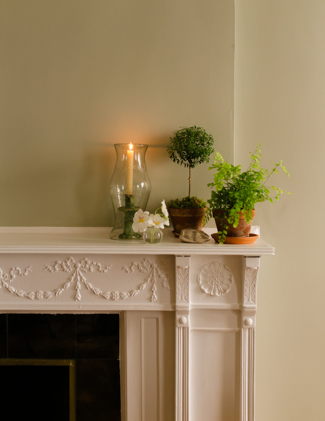 A styled interior scene showing a white ornate fireplace mantel with potted green plants, a lit glass candle holder, and small white flowers against a warm beige wall.