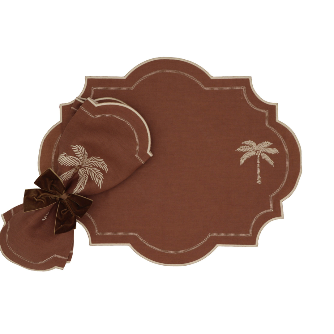 A brown placemat set with scalloped edges, featuring a palm tree motif and a matching napkin tied with a dark brown bow, shown on a plain white background.