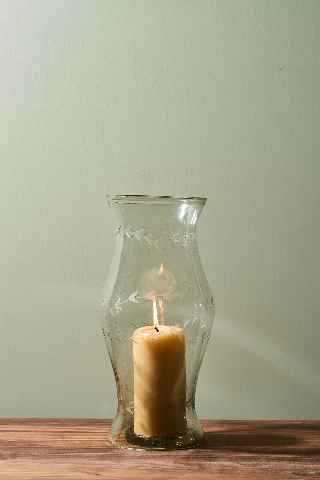 A lit cream-colored pillar candle inside a clear glass hurricane lantern with etched floral patterns, set on a wooden surface against a pale sage green wall.