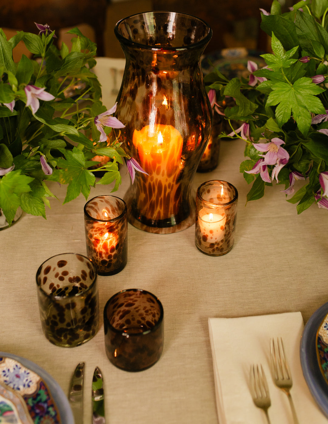 A set of amber glass candle holders and a matching vase with lit candles, styled on a beige table runner with green foliage and purple flowers.