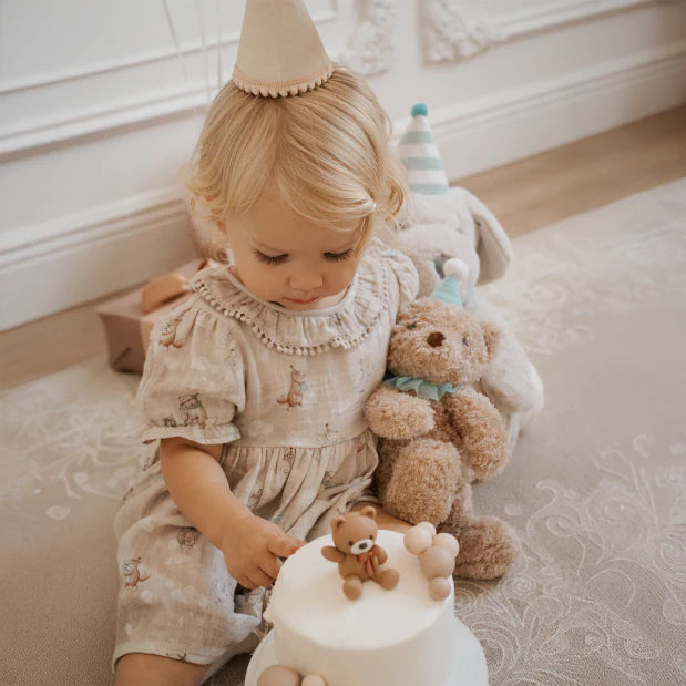 Child in a light dress with teddy bears and a party hat on a neutral background
