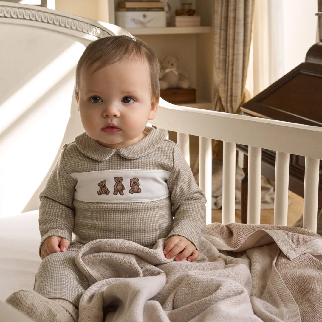 Baby in a checkered outfit with teddy bear design sitting in a crib.